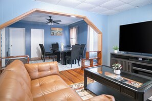 Un salon avec un canapé en cuir marron, un coin repas avec des chaises, un téléviseur et un ventilateur de plafond.