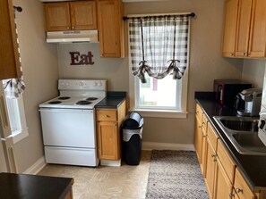 A kitchen with wooden cabinets, a white stove, a trash can, and a window with plaid curtains. - Cozy Upstairs 2BR Apartment Near Hospitals & Charleston (Saint Albans)