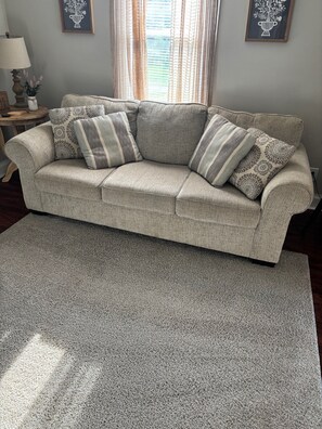 A beige sofa with patterned cushions, a grey textured rug, and framed wall art. - Cozy Upstairs 2BR Apartment Near Hospitals & Charleston (Saint Albans)