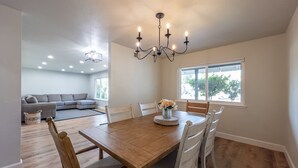 A dining area with a wooden table, white chairs, a chandelier, and a view of a living room with a sofa and a window.