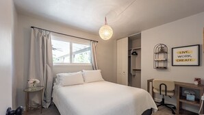 A bedroom with a bed, a desk, a chair, a hanging light fixture, and a window with curtains.