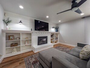 A modern living room with a fireplace, a flat-screen TV mounted above it, and a ceiling fan.