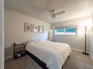 A bedroom with a bed, a nightstand, a ceiling fan, framed pictures, a window with a view, and a floor lamp.