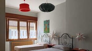 Quarto com duas camas, radiador, janela com cortinas, ventoinha no teto e luminária na parede.