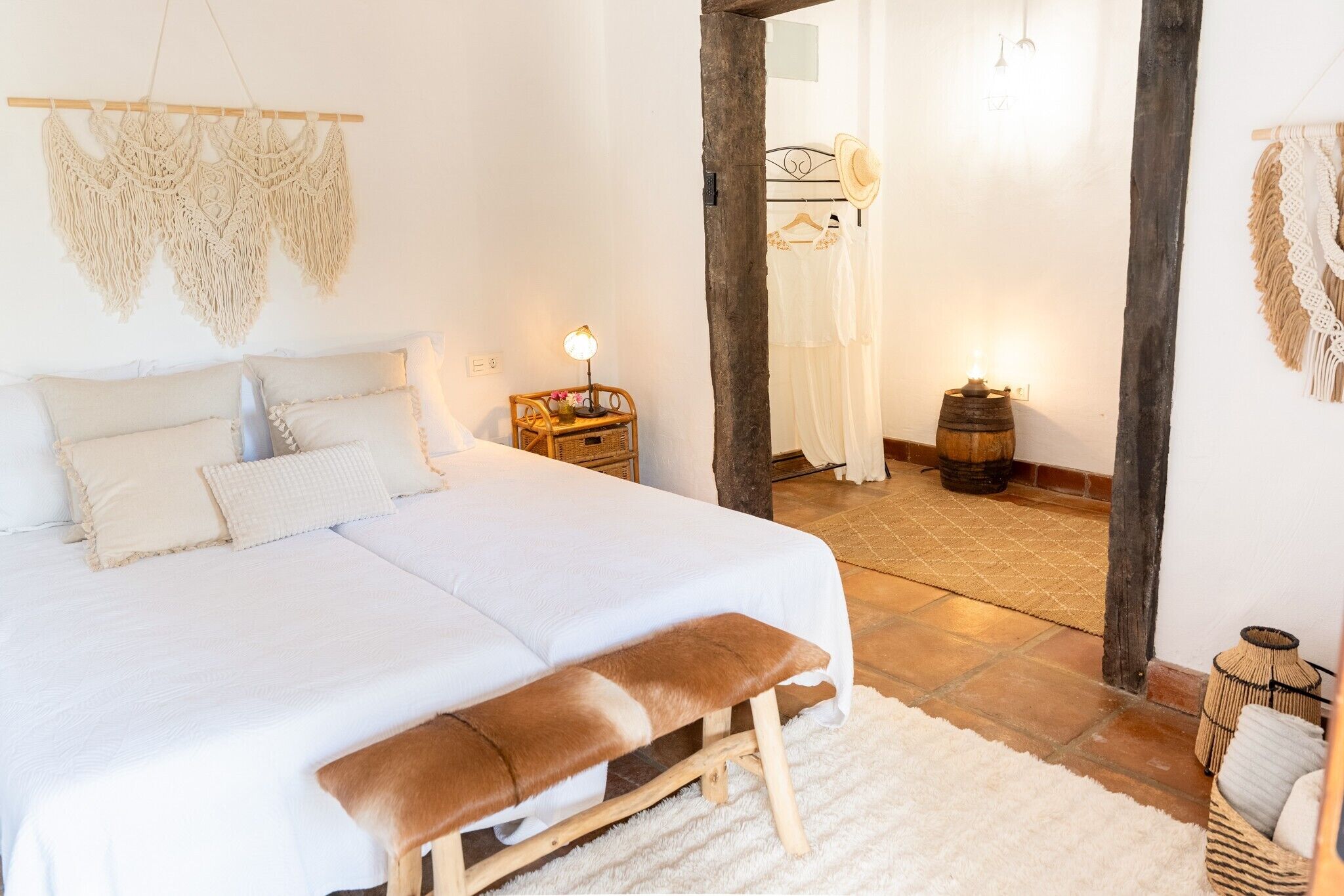 A bedroom with a large bed, a wooden bench, macramé wall hangings, a nightstand with a lamp, and a rustic barrel lamp.