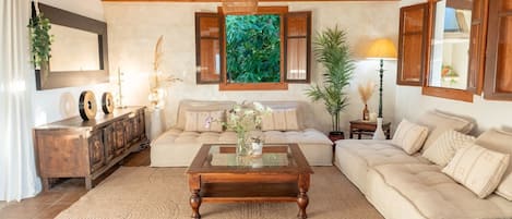 A cozy living room with a wooden ceiling, a glass coffee table, a sofa, and a large window with a view of greenery.