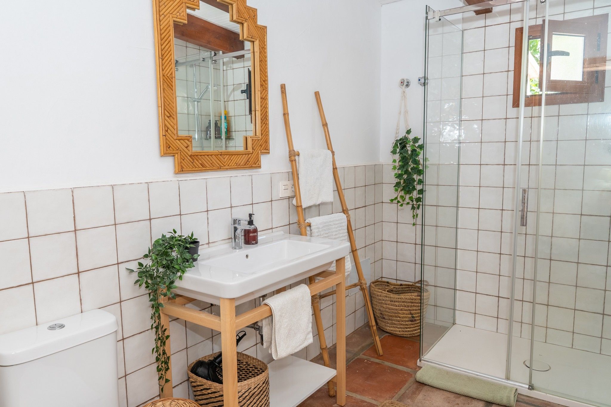 Een badkamer met een wit wastafelmeubel, spiegel en douche. Er staan planten en manden in de buurt.