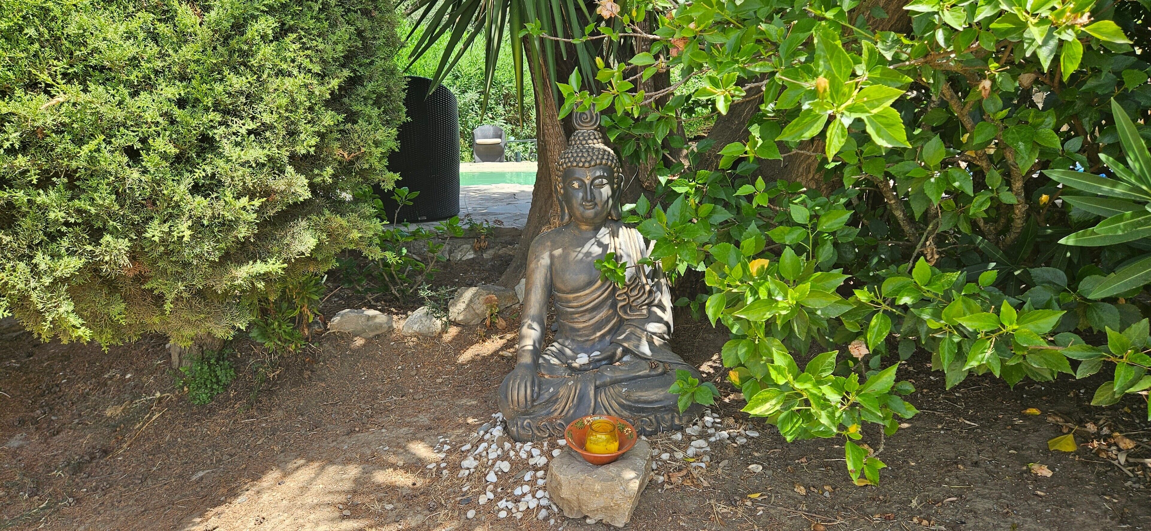 Una estatua de una figura sentada en un jardín con follaje circundante y un pequeño recipiente de ofrendas.
