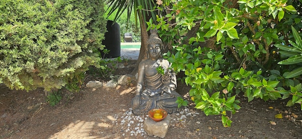 A statue of a seated figure in a garden with surrounding greenery and a small bowl of offerings.