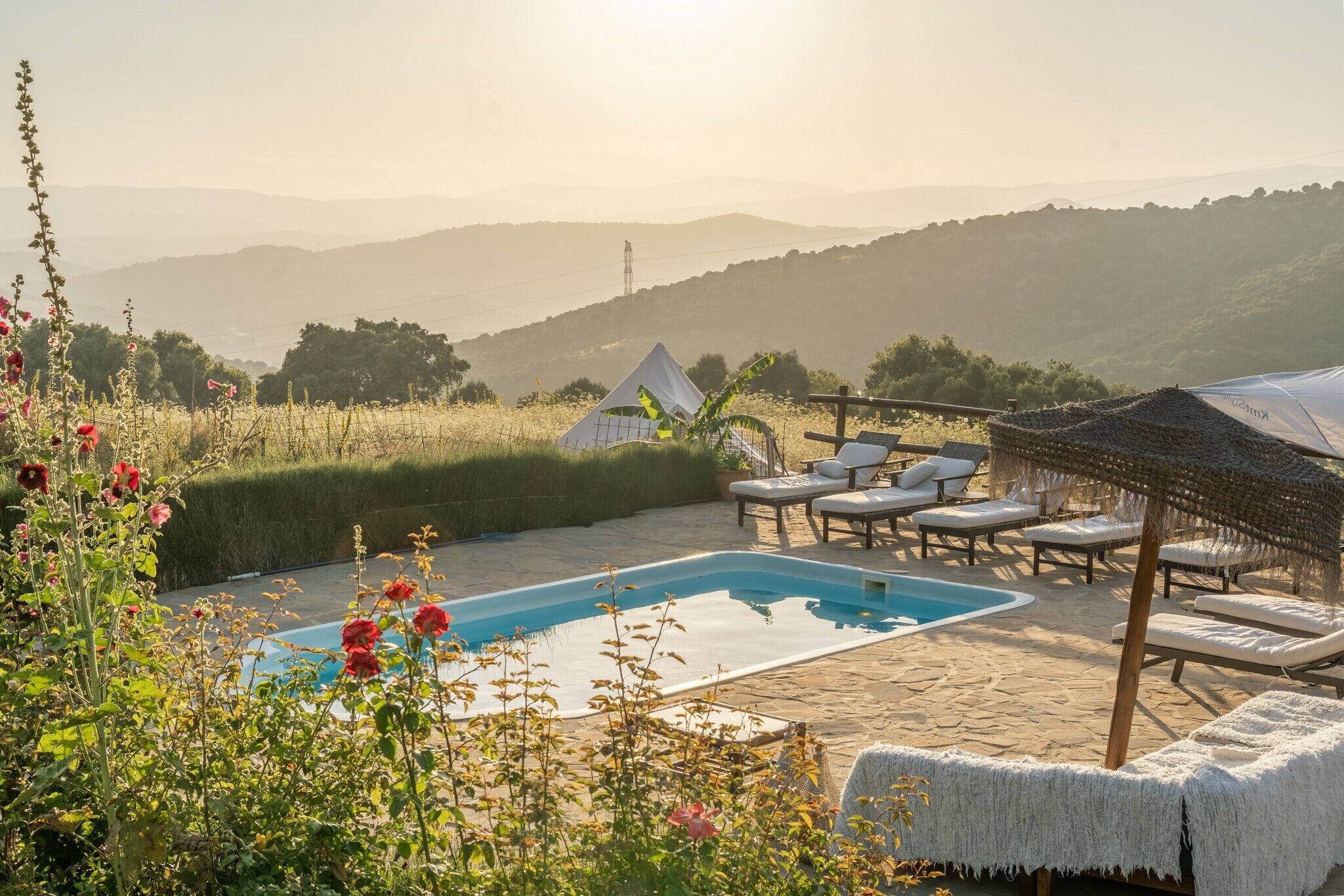 A pool area with lounge chairs and a tent, surrounded by greenery and hills.