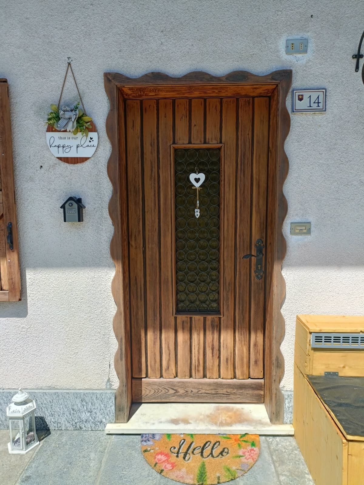 Una puerta de madera con un ventanal en forma de corazón, un felpudo con la palabra 'Hello' y un diseño floral, y una placa con el número 14 en la pared.