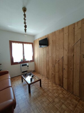 A room with a wooden wall, a television mounted on it, a window with curtains, a table with a vase, and a ceiling fan.