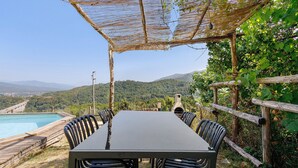 Uma área de refeições ao ar livre com mesa e cadeiras, uma pérgola e vista para uma piscina e montanhas.
