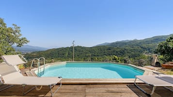 Une piscine d’un bleu éclatant, bordée de chaises longues sur une terrasse en bois, le tout entouré d’une végétation luxuriante et de collines.