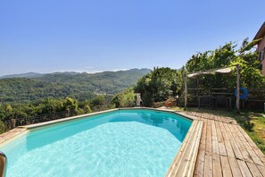 A rectangular swimming pool with a wooden deck surrounding it, set against a backdrop of a mountainous landscape.