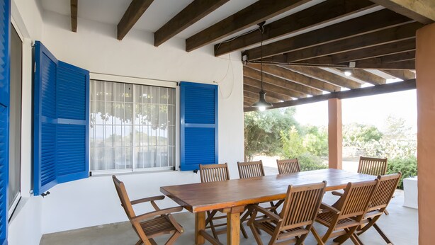 A covered outdoor dining area with a wooden table and chairs, a window with blue shutters, and a view of greenery outside.