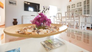 A room with a glass table displaying seashells and a magazine, a white cabinet with glass doors, and a television on the wall. - Casa Rita in Formentera with Sea Views (Sant Ferran de ses Roques)