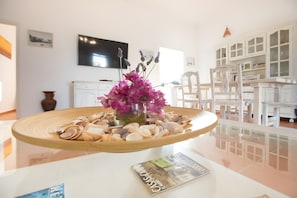 A room with a glass table displaying seashells and a magazine, a white cabinet with glass doors, and a television on the wall. - Casa Rita in Formentera with Sea Views (Sant Ferran de ses Roques)
