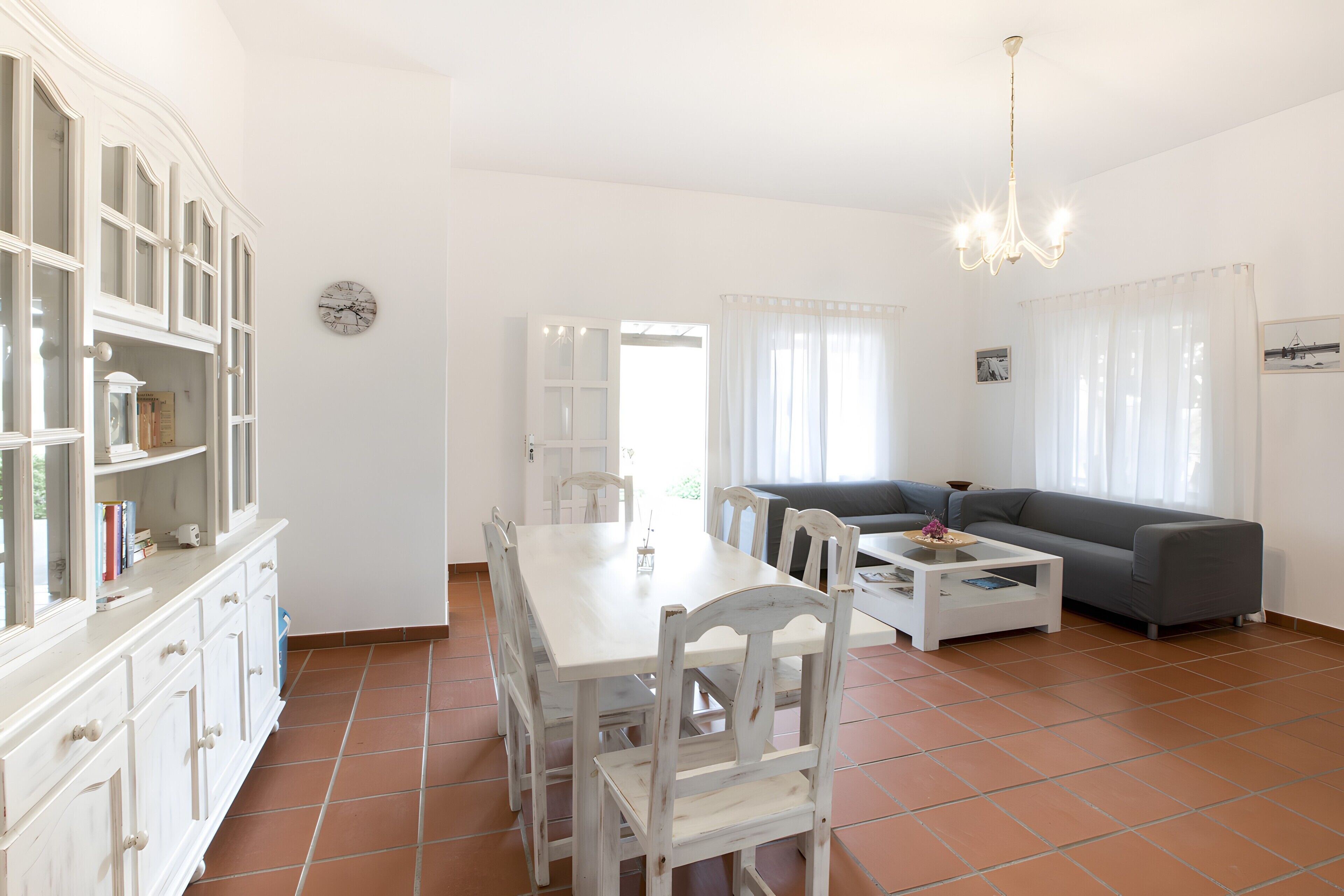 Amplia sala de estar con mesa de comedor, sillas, sofá y una mesa de centro. Hay una estantería y un reloj en la pared.