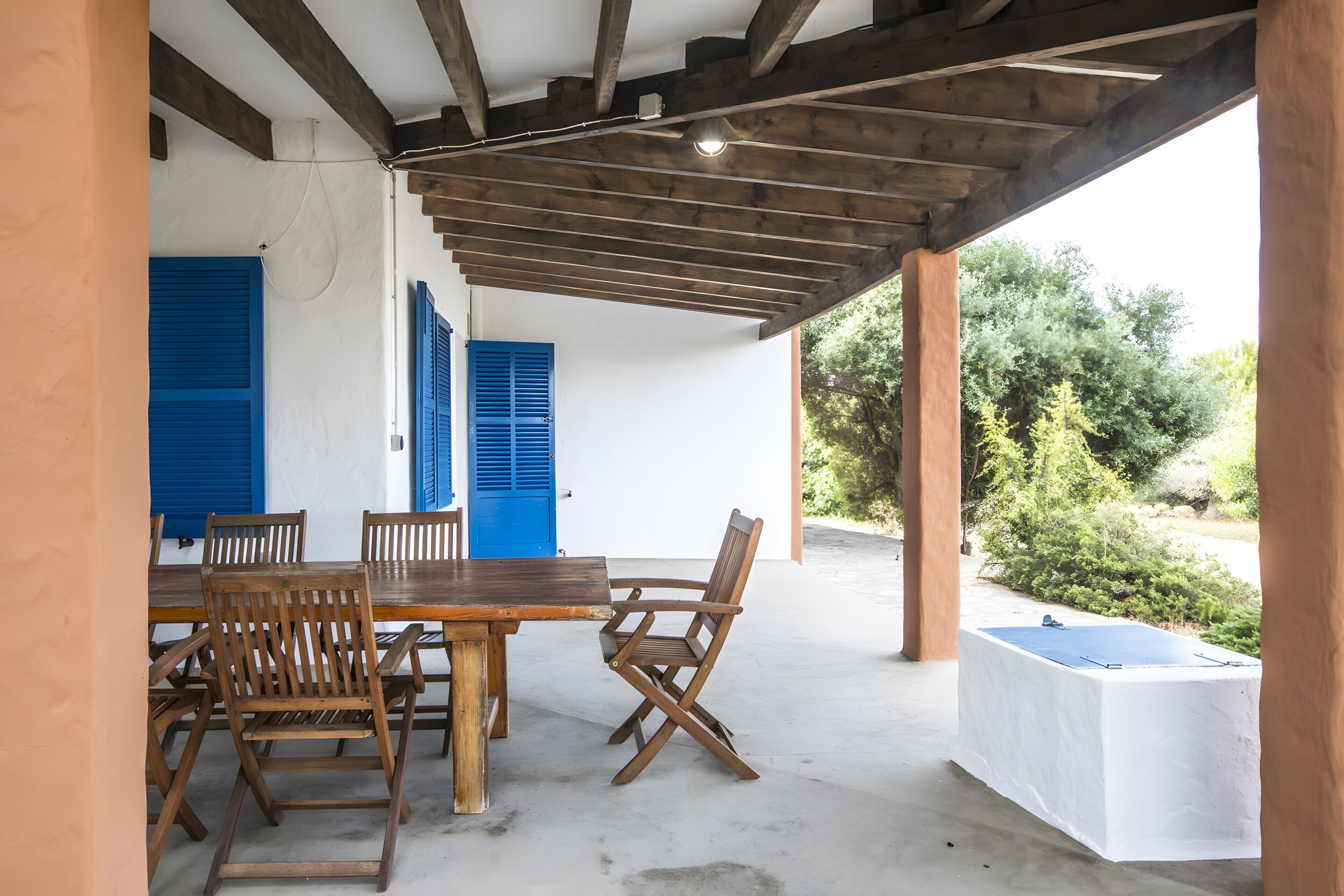 Un patio al aire libre con una mesa y sillas de madera, contraventanas azules y una pared blanca.