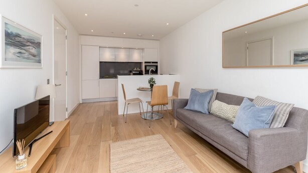 Een moderne woonkamer met een grijze bank, een televisie en een eethoek met stoelen en een tafel.