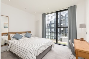 A modern hotel room with a large bed, a desk, and a view of the city through floor-to-ceiling windows. - Charming 2 bedroom apartment in Quartermile (Edinburgh)