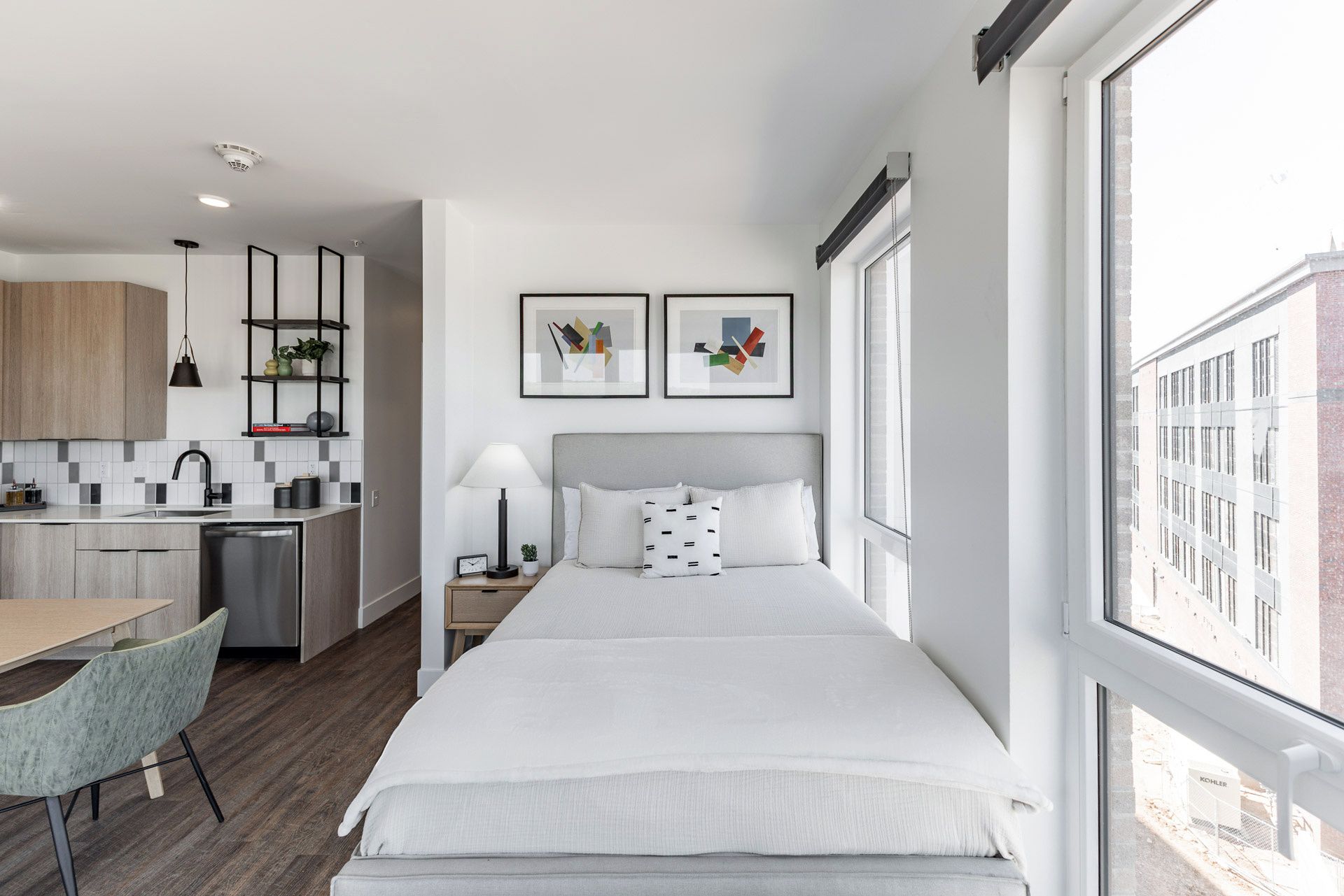 Ein kleines Hotelzimmer mit Bett, Nachttisch, einem Schreibtisch mit Stuhl, einer Küchenzeile und einem großen Fenster mit Blick auf die Stadt.