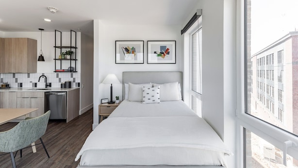 Ein kleines Hotelzimmer mit Bett, Nachttisch, einem Schreibtisch mit Stuhl, einer Küchenzeile und einem großen Fenster mit Blick auf die Stadt.