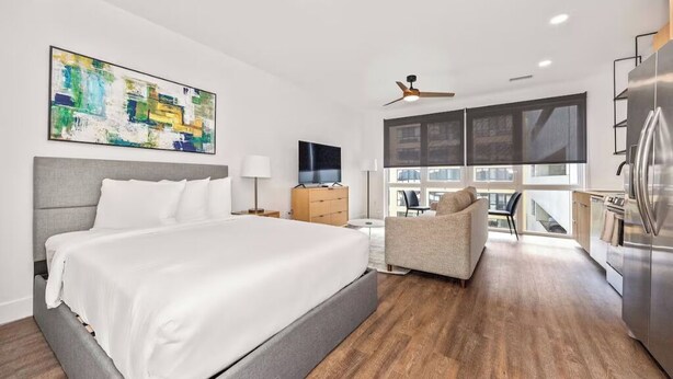 Ein modernes Hotelzimmer mit einem großen Bett, einer Couch, einem Fernseher, einem Deckenventilator und Blick auf die Stadt durch große Fenster.