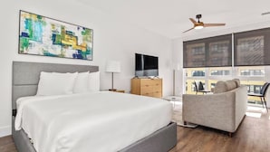 Ein modernes Hotelzimmer mit einem großen Bett, einem Flachbildfernseher, einem Deckenventilator und Blick auf die Stadt durch große Fenster.