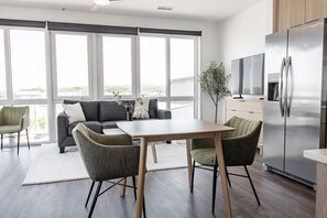 Una cocina moderna con un refrigerador de acero inoxidable, una mesa de comedor con sillas y un sofá con vistas a través de grandes ventanales.