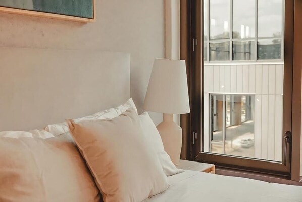 Quarto de hotel com cama, almofadas, luminária e janela com vista para edifícios.