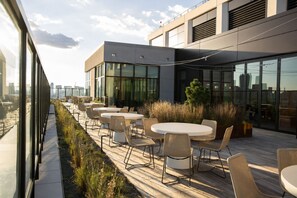 Terraço ao ar livre com mesas e cadeiras redondas, deck de madeira e vista para o horizonte urbano.