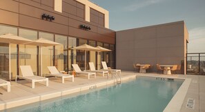 Uma área de piscina no terraço com espreguiçadeiras e guarda-sóis, piscina com água azul cristalina e fachada de um edifício moderno.