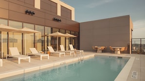 Zona de piscina en la azotea con sillones de descanso y sombrillas, piscina con agua cristalina y fachada de un edificio moderno.