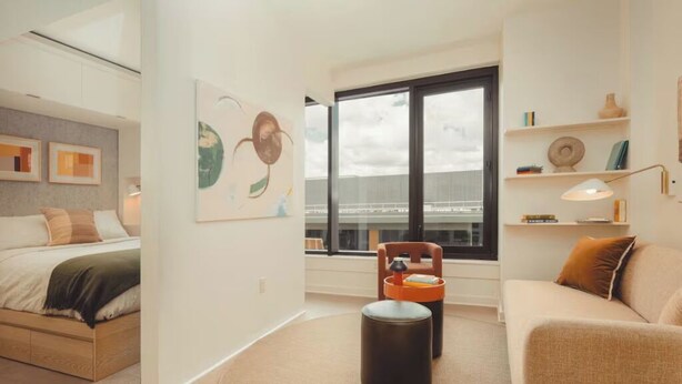 A modern bedroom with a bed, a round ottoman, a sofa, and a window with a view of a building. - Premium Studio Low Floor w/ Kitchen, Heated Rooftop Pool & Gym (Atlanta)