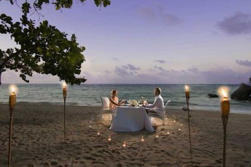 A beachside dining setup with a table and chairs under a tree, surrounded by candles.