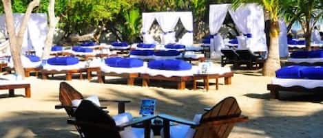 A beachside lounge area with lounge chairs and tables, surrounded by palm trees and a sandy beach.