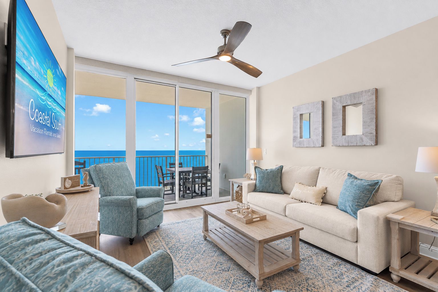 Un salon spacieux avec un balcon donnant sur la mer, un ventilateur de plafond et un grand écran de télévision.