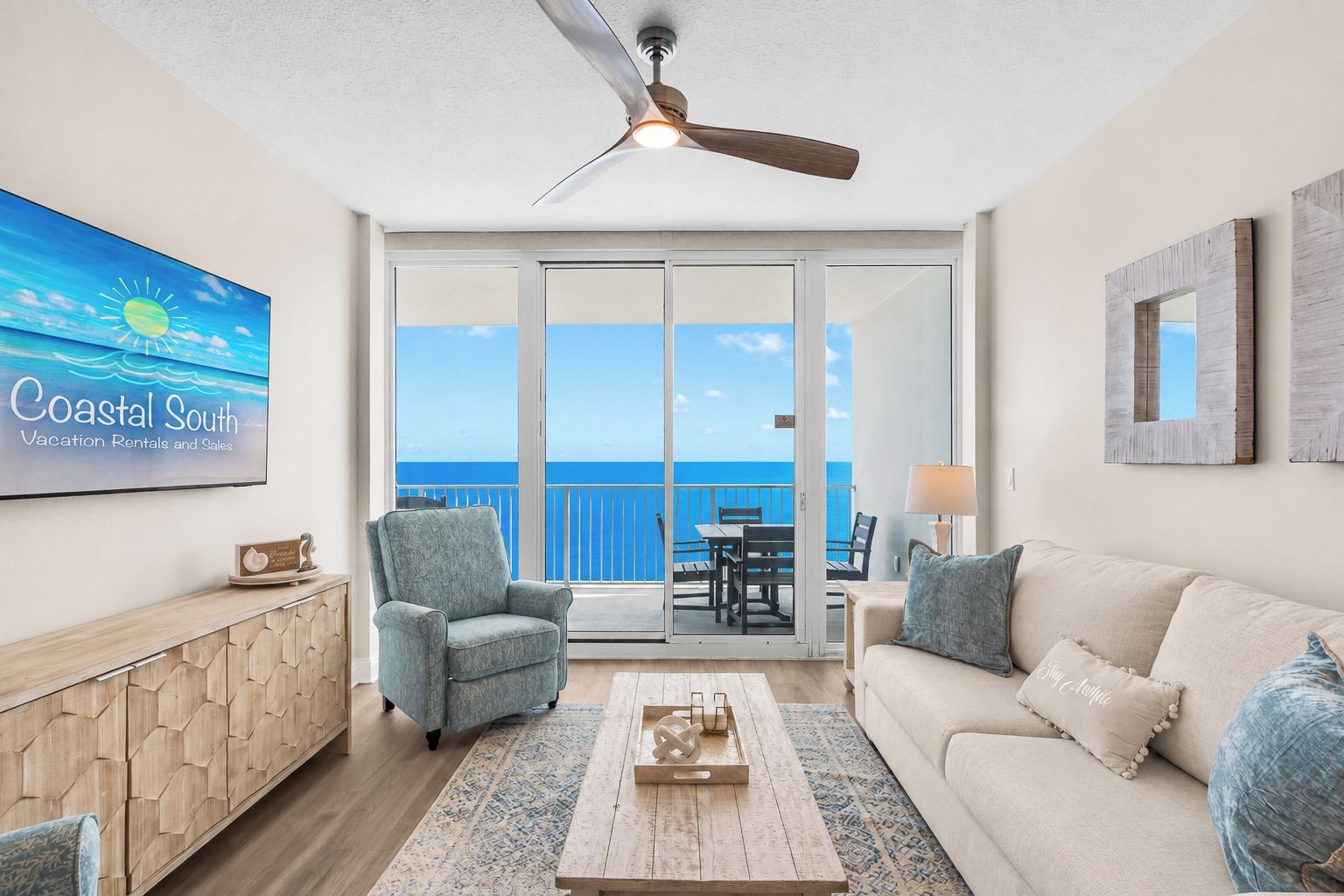 A spacious living room with a view of the ocean, a ceiling fan, a flat-screen TV, and a dining area with a table and chairs.