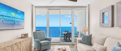 A spacious living room with a view of the ocean, a ceiling fan, a flat-screen TV, and a dining area with a table and chairs.
