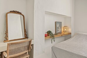 A room with a large ornate mirror, a wooden chair, a shelf with decorative items, and a bed with a textured cover.