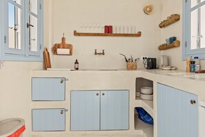 A kitchen with blue cabinets, a wooden cutting board, a hanging rack with glasses, and a coffee machine. - Maison Pnoé – Ihr Ferienhaus im Herzen von Arkasa mit Meer- und Sonnenuntergangsblick (Αρκάσα)