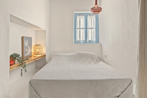 Une pièce comprenant un lit, une fenêtre avec des volets bleus, un luminaire suspendu et une étagère fixée au mur, ornée d’objets décoratifs.