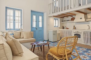 A cozy living room with a sofa, a wooden chair, a small table, and a kitchen area with a refrigerator, oven, and shelves.