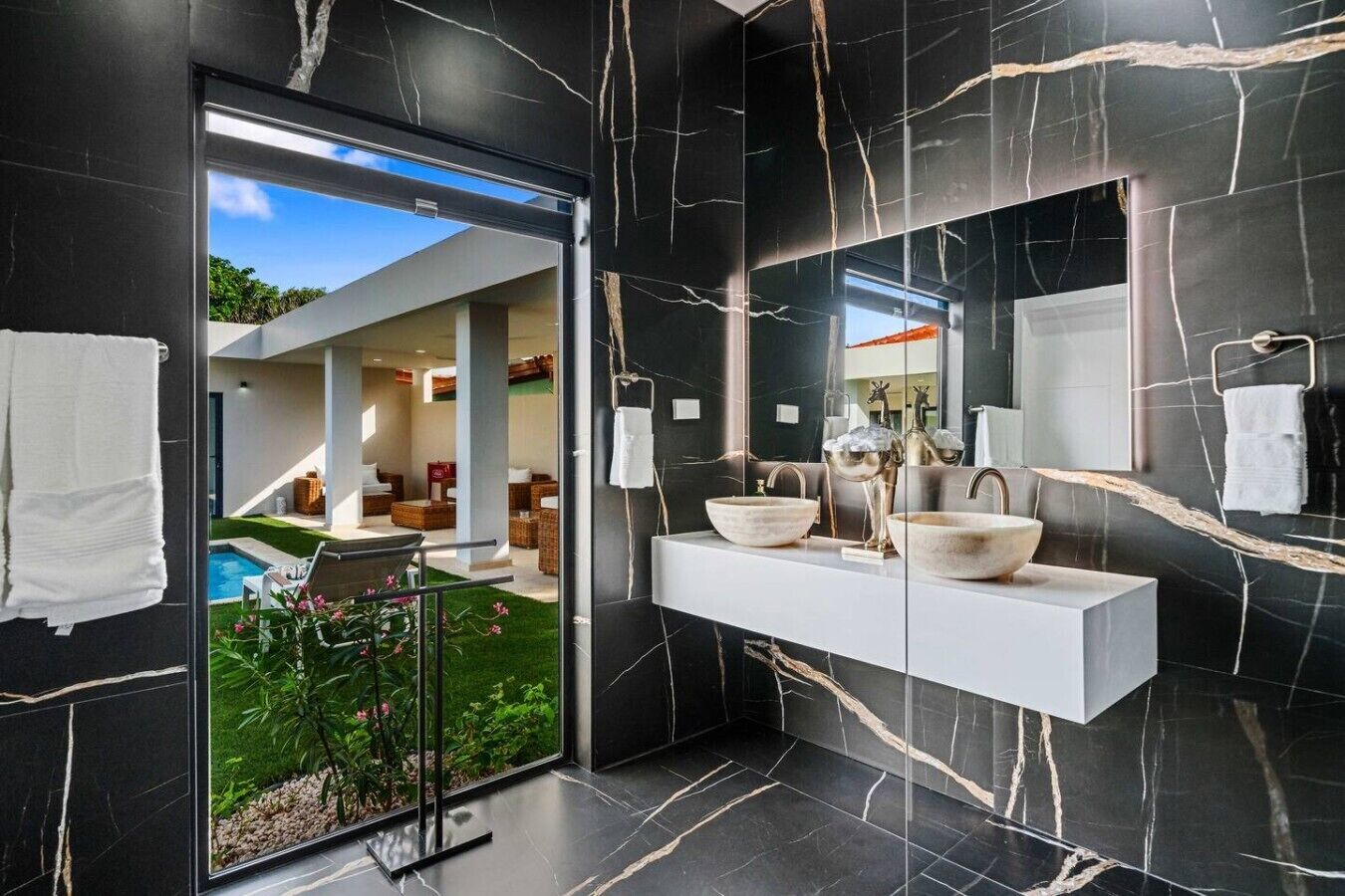 Un baño moderno con un espejo grande, dos lavamanos y una puerta de cristal que da a una zona de piscina.