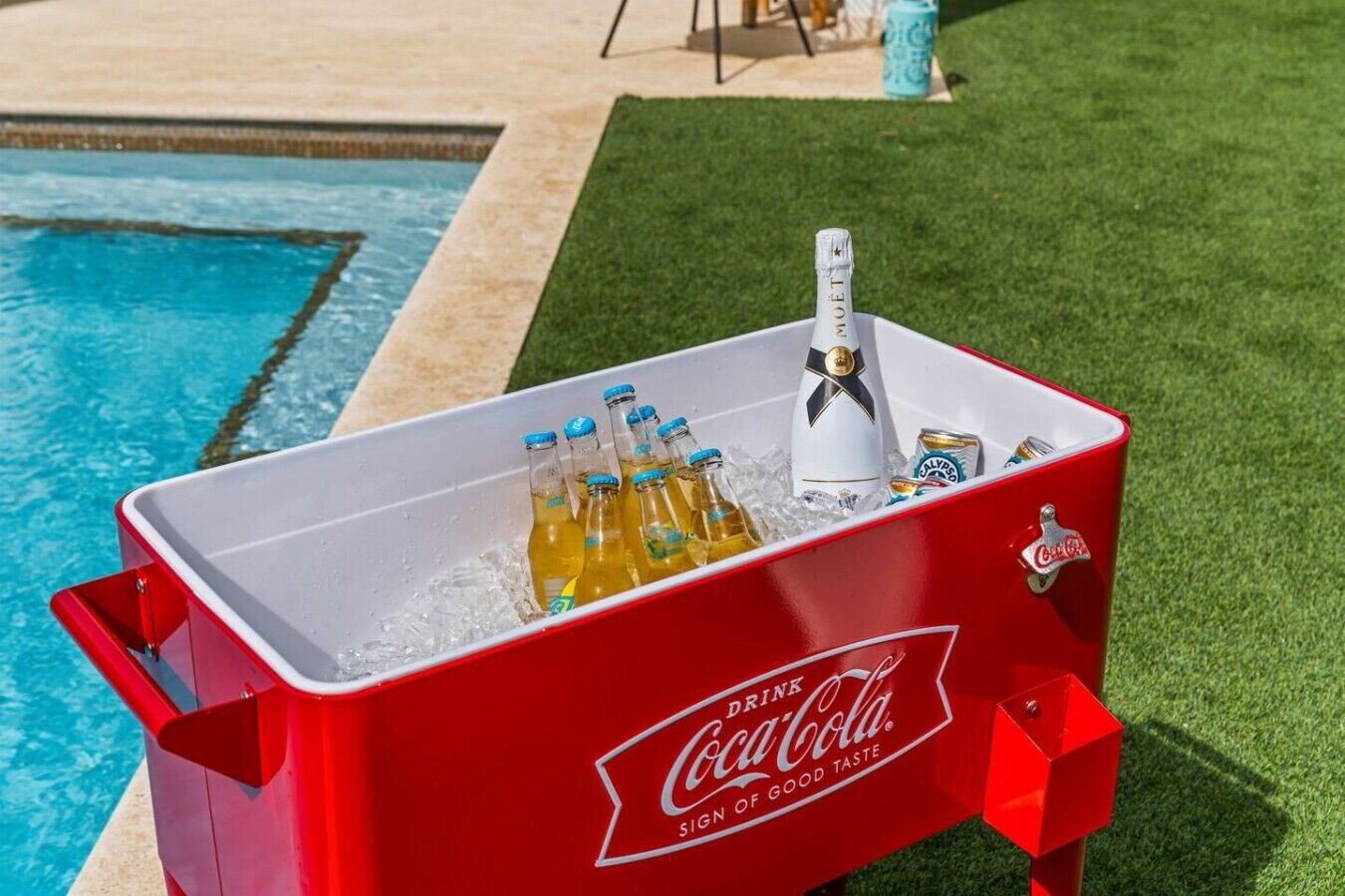 A red Coca-Cola branded cooler filled with bottles and a champagne bottle, placed near a swimming pool.