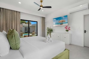 Ein Schlafzimmer mit Bett, Deckenventilator, Fernseher, einem Fenster mit Vorhängen und Blick auf einen Außenpoolbereich.