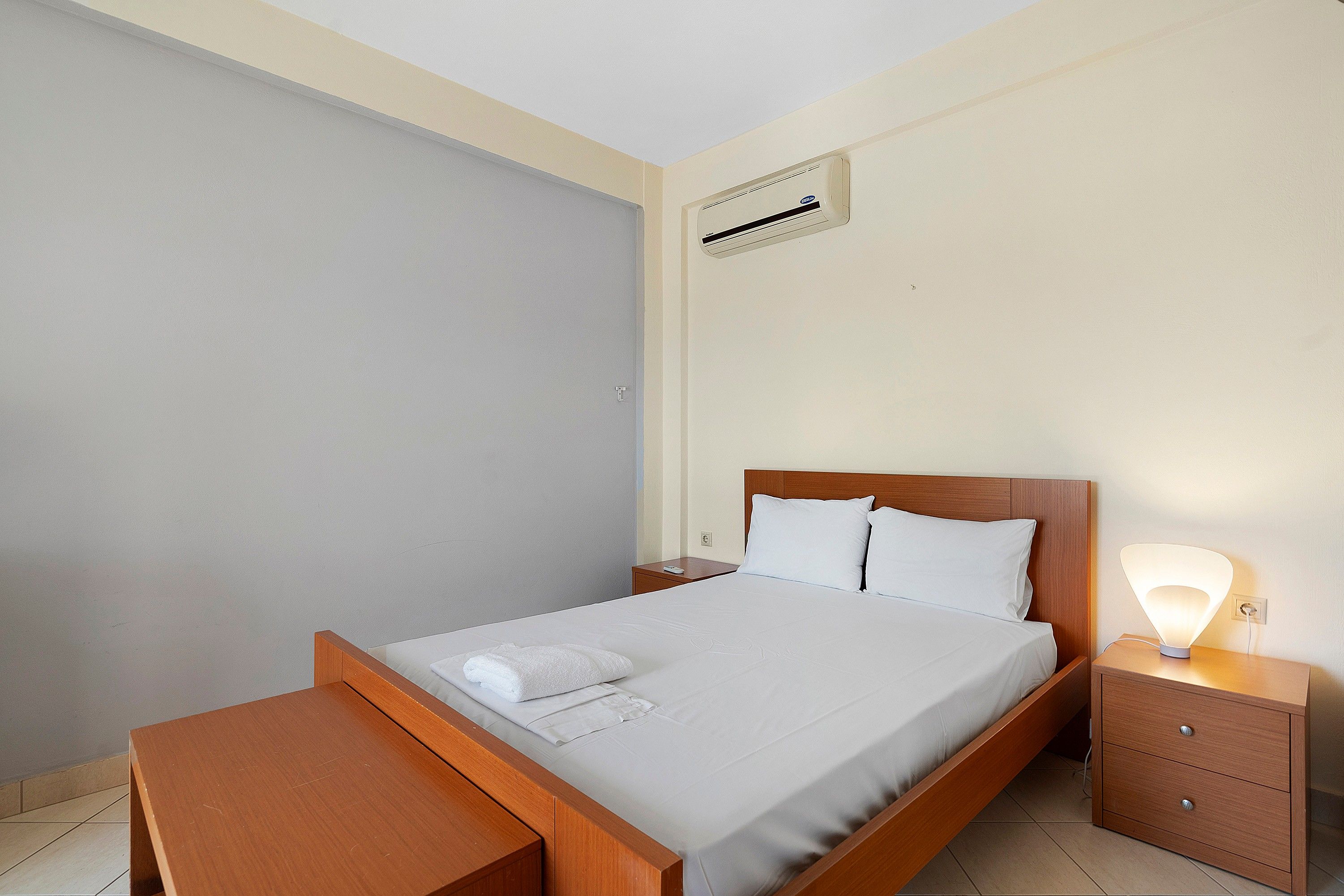 A hotel room with a wooden bed, white linens, a bedside lamp, and a wall-mounted air conditioner.