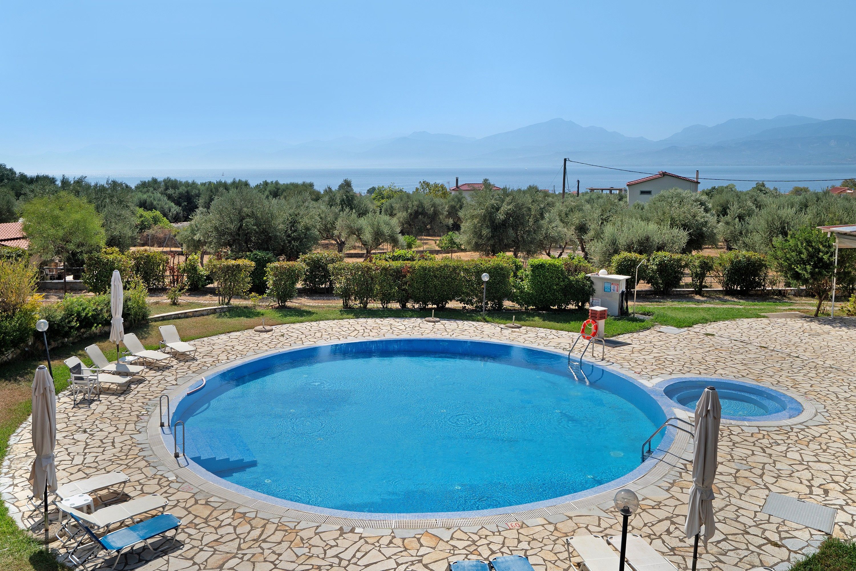 Una piscina con sillas de descanso y sombrillas, rodeada de baldosas de piedra, con vistas al mar y montañas al fondo.